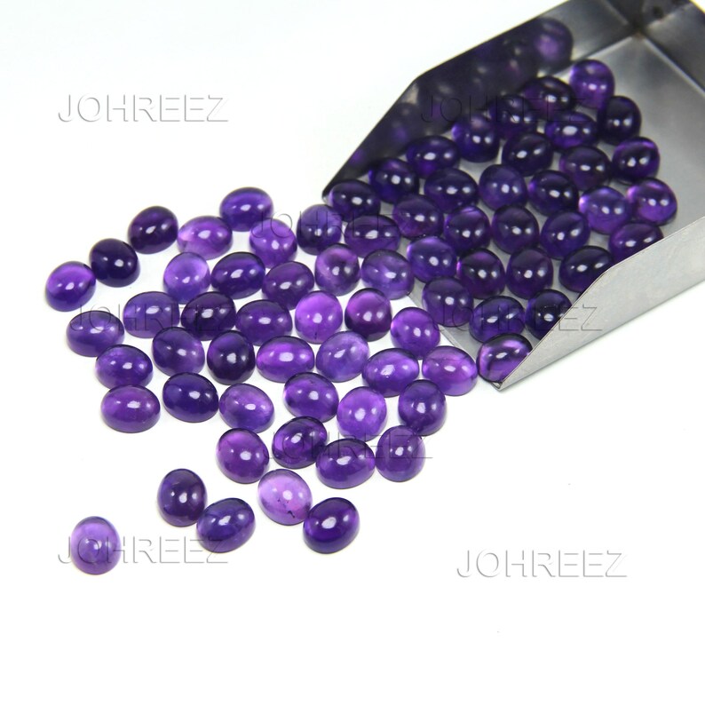 May include: A collection of oval-shaped, polished amethyst gemstones in varying shades of purple. The gemstones are scattered on a white surface, with some spilling from a metal scoop. The text "JOHREEZ" is visible.