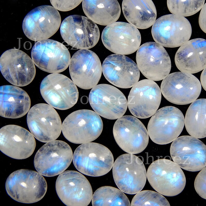 May include: A collection of oval-shaped moonstone cabochons, each displaying a unique iridescent blue sheen. The stones are arranged on a black background.