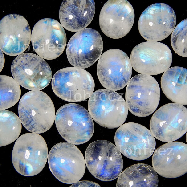 May include: A collection of oval-shaped moonstone cabochons, each with a milky white base and iridescent blue and green flashes. The stones are arranged in a random pattern on a black background.