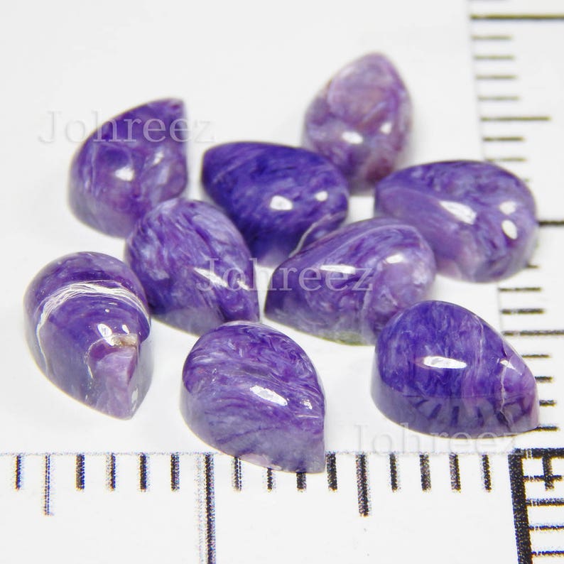 May include: A collection of eight purple and white teardrop-shaped cabochons. The cabochons are smooth and polished, with a swirling pattern of purple and white.
