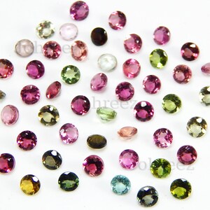May include: Assortment of round, faceted gemstones in various colours, including pink, green, blue, and brown. The gemstones are arranged on a white surface.