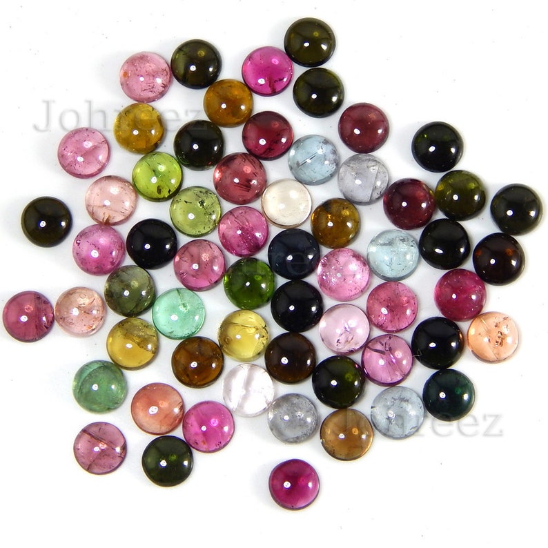 May include: A collection of round, multi-colored gemstones. The gemstones are various shades of pink, green, brown, blue, and black. The gemstones are scattered on a white surface.