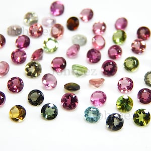 May include: Assortment of round, faceted gemstones in various colours, including pink, green, blue, and brown. The gemstones are loose and scattered on a white surface.