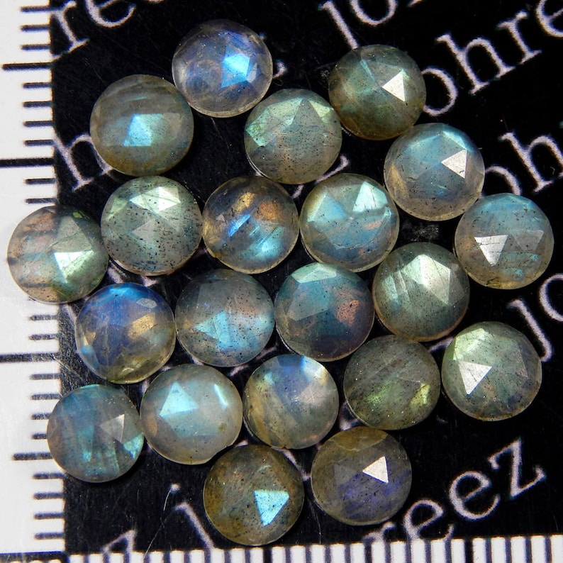 May include: A collection of 20 iridescent green and blue labradorite gemstones, each with a faceted round shape. The stones are arranged in a random pattern on a black surface.