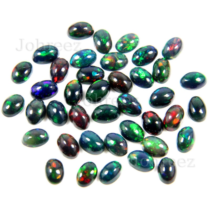 May include: A collection of oval-shaped black opals with green, red, and blue iridescent flashes. The opals are loose and scattered on a white background.
