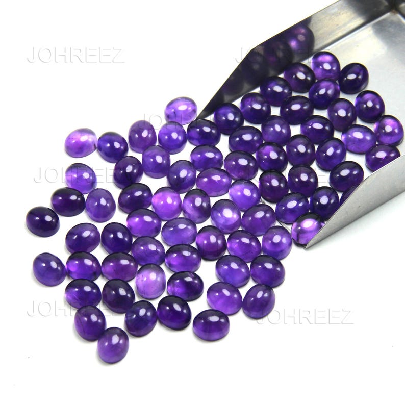 May include: A collection of oval-shaped, polished amethyst gemstones in varying shades of purple. The gemstones are displayed in a metal scoop, with some scattered on a white surface. The gemstones have a smooth, glossy finish.