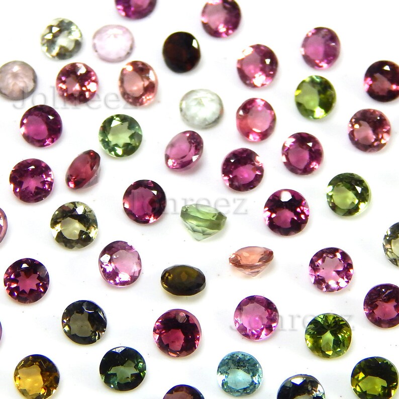 May include: Assortment of round, faceted gemstones in various colours, including pink, green, brown, and clear. The gemstones are arranged on a white surface.