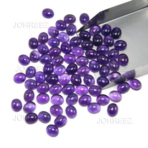 May include: A collection of oval-shaped, polished amethyst gemstones in varying shades of purple. The gemstones are scattered on a white surface, with a metal tool partially visible in the upper right corner. The gemstones are ready for jewelry making.