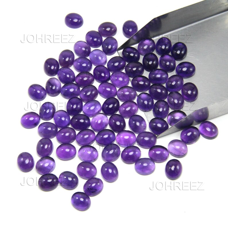 May include: A collection of oval-shaped, polished amethyst gemstones in varying shades of purple. The gemstones are scattered on a white surface, with a metal tool partially visible in the upper right corner. The gemstones are ready for jewelry making.