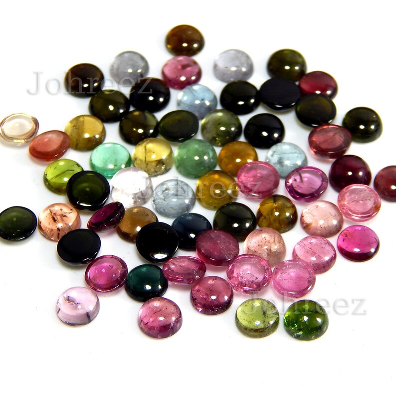 May include: A collection of round, cabochon-cut gemstones in various colors, including pink, green, blue, and brown. The gemstones are arranged in a loose pile on a white background.