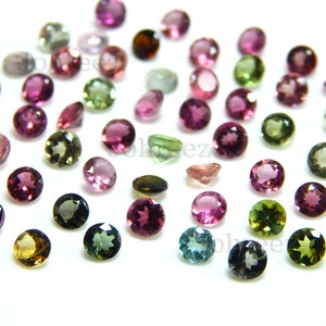 May include: Assortment of round, faceted gemstones in various colours, including pink, green, blue, and brown. The gemstones are scattered on a white surface.