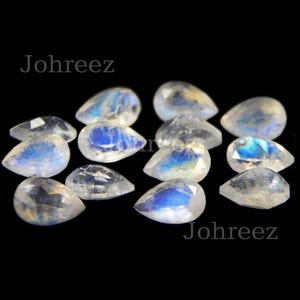 May include: A collection of pear-shaped moonstone gemstones with iridescent blue and white colors. The stones are arranged on a black background.