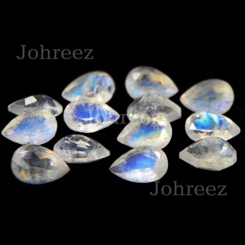 May include: A collection of pear-shaped moonstone gemstones with iridescent blue and white colors. The stones are arranged on a black background.