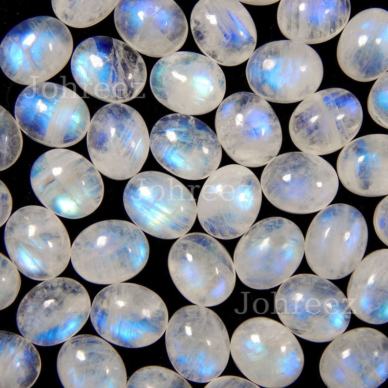 May include: A collection of oval-shaped moonstone cabochons with a white iridescent sheen and blue flashes. The stones are arranged in a random pattern on a black background.