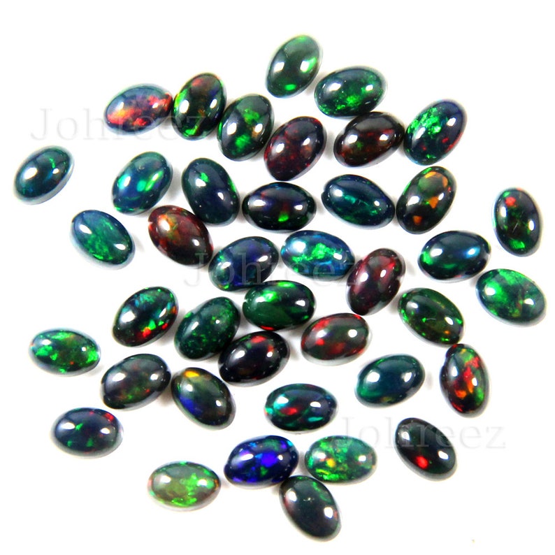May include: A collection of oval-shaped black opals with green, red, and blue iridescent flashes. The opals are scattered on a white surface.