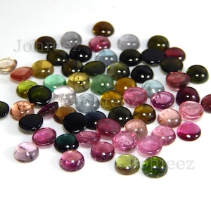 May include: A collection of round, multi-colored gemstones. The gemstones are various shades of pink, green, brown, blue, and black. The gemstones are arranged in a loose pile on a white surface.