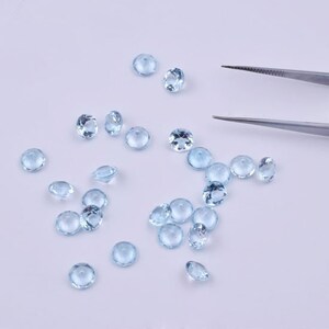 Pode incluir: Uma cole&ccedil;&atilde;o de pedras preciosas redondas de cor azul claro, espalhadas sobre uma superf&iacute;cie branca. As pedras preciosas s&atilde;o de v&aacute;rios tamanhos e parecem ser facetadas.
