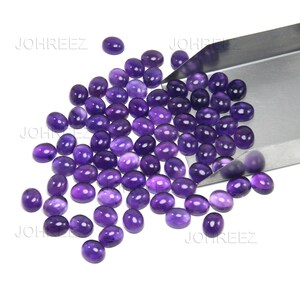 May include: A collection of oval-shaped, polished purple gemstones. The gemstones are scattered across a white surface, with some held in a silver metal tray. The gemstones have a smooth, glossy finish and vary slightly in shade.
