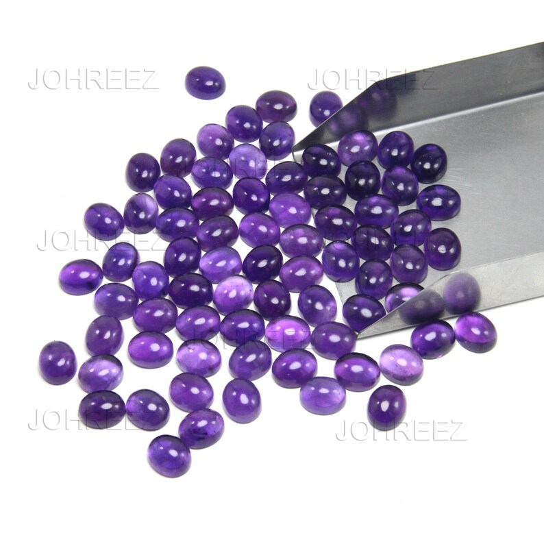 May include: A collection of oval-shaped, polished purple gemstones. The gemstones are scattered across a white surface, with some held in a silver metal tray. The gemstones have a smooth, glossy finish and vary slightly in shade.