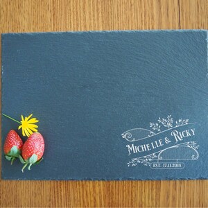 May include: A black slate serving board with a white engraved design that reads "Michelle & Ricky Est. 17.11.2018". Two red strawberries and a yellow flower are on the board.