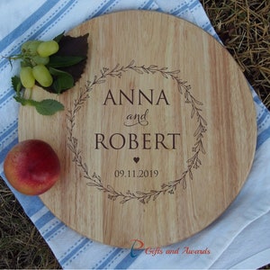 May include: A round wooden serving board with a floral wreath design engraved on the surface. The wreath encloses the names "Anna and Robert" and the date "09.11.2019".