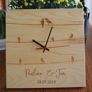 May include: A wooden clock with a red second hand and black hour and minute hands. The clock face has a design of birds on a wire. The clock is personalised with the names "Pauline & Tim" and the date "28.09.2018".