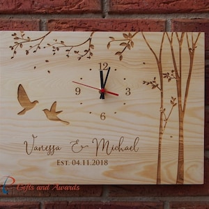 May include: A wooden clock featuring a tree and bird design. The clock face has black hands and Roman numerals. The clock is personalised with the names "Vanessa & Michael" and the date "EST. 04.11.2018".