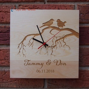 May include: A wooden clock featuring a carved tree branch design with two birds perched on the branch. The clock face has black hands and a red second hand. The clock is personalised with the names "Tammy & Don" and the date "06.11.2018".