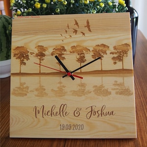 May include: A square wooden clock with a laser-etched landscape scene featuring trees, birds, and a mirrored reflection. The clock hands are black with a red second hand. The names "Michelle & Joshua" and the date "19.09.2020" are also etched.