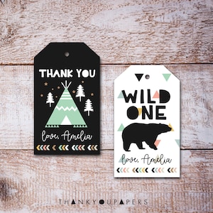 Puede incluir: Dos etiquetas de regalo en blanco y negro con un tema de bosque. La primera etiqueta dice "Gracias" con una tienda de campaña y árboles. La segunda etiqueta dice "Salvaje Uno" con un oso y formas geométricas. Ambas etiquetas tienen el texto "amor, Amelia" y una fila de flechas en la parte inferior.