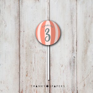 May include: A pink and white striped lollipop with the text "Turning 3" on it. The lollipop is on a white stick against a white wood background.