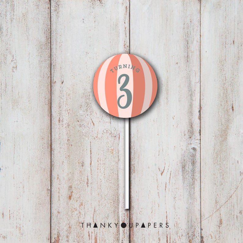 May include: A pink and white striped lollipop with the text "Turning 3" on it. The lollipop is on a white stick against a white wood background.