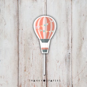 May include: A hot air balloon cupcake topper with a red and white striped balloon, a gray basket, and a string of colorful flags. The topper is on a white stick.