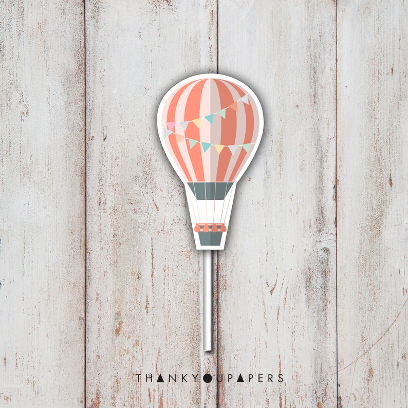 May include: A hot air balloon cupcake topper with a red and white striped balloon, a gray basket, and a string of colorful flags. The topper is on a white stick.