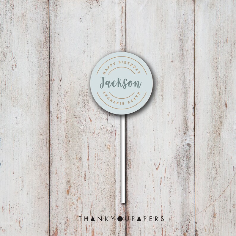 May include: A light blue round cake topper with the text "Happy Birthday" in a gold circle around the text "Jackson". The cake topper is on a white stick against a wood background.
