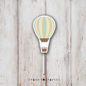 May include: A light blue and yellow hot air balloon with a brown basket, on a white stick. The balloon has a white background with yellow stripes and a light blue center. The basket has a brown rope and a brown handle. The stick is white and has a white background. The image is on a white wooden background.