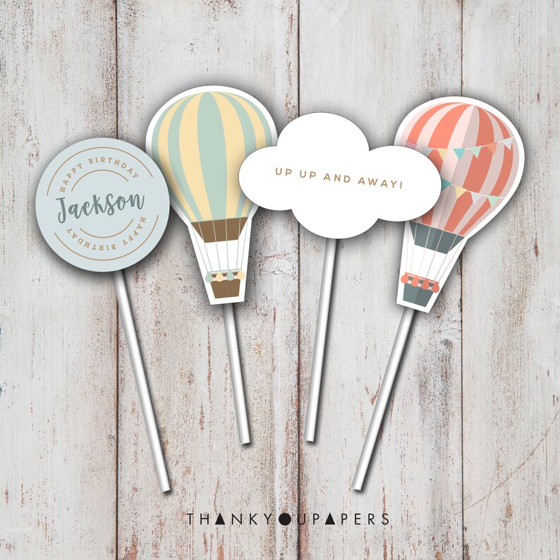 May include: Three hot air balloon themed cake toppers. One topper is a circle with the text "Happy Birthday Jackson Happy Birthday". Another topper is a white cloud with the text "Up Up And Away!" The third topper is a hot air balloon with red and white stripes and a banner of flags.