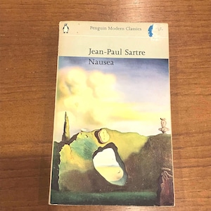May include: A copy of "Nausea" by Jean-Paul Sartre, a Penguin Modern Classics paperback. The cover art depicts a surreal landscape with a clock and a statue. The book is resting on a wooden surface.
