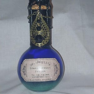 Puede incluir: Una botella de poción azul con una etiqueta que dice "Dr. Jekyll's Transforming Potion". La botella tiene un sello de cera negra y un dije de filigrana dorada. El color de la poción cambia de azul a verde.