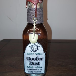 Pode incluir: Uma garrafa de vidro marrom com um selo de cera e um rótulo que diz "Goofer Dust Guaranteed to be locally sourced". O rótulo também apresenta um pentagrama e uma pequena pá.