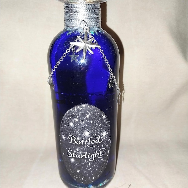 May include: A cobalt blue glass bottle with a silver-colored cap and a decorative chain of stars. The bottle has a label that reads "Bottled Starlight" with a starry background. The bottle is approximately 8 inches tall.