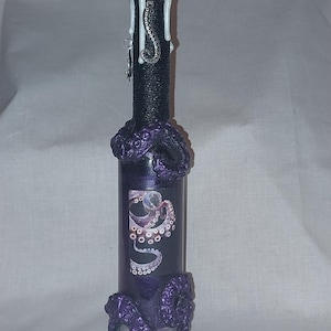 May include: A decorative glass bottle with a black wrapped neck and a cork stopper covered in white wax. The bottle is adorned with purple octopus tentacles and a graphic of an octopus. A small silver charm hangs from the neck.