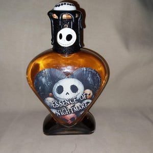 May include: A heart-shaped, amber-colored glass bottle with a black base and stopper. The bottle features a label with a skull design and the text "Essence of Nightmare." The stopper is decorated with a skull and dripping black wax.