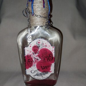 May include: A clear glass bottle with a label reading "Sinners Corn Liquor" and a decorative top. The top is wrapped in twine and adorned with blue and red wax, and a silver chain with charms. The bottle contains red sand.