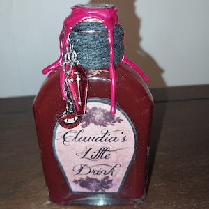 May include: A small glass bottle with a red liquid inside. The bottle is sealed with a pink wax seal and has a black string tied around the neck. A silver charm with a red vampire mouth and a coffin hangs from the string. The label on the bottle reads "Claudia's Little Drink".