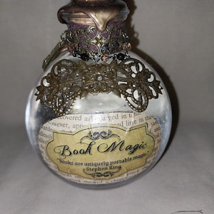 May include: A clear, round glass bottle with a decorative metal filigree and a bronze-colored stopper. The bottle has a label that reads "Book Magic" with a quote by Stephen King. The bottle is filled with a clear liquid and paper.