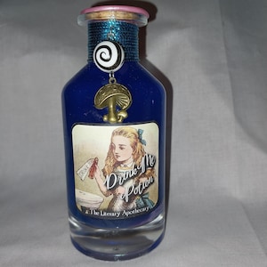 May include: A blue potion bottle with a cork stopper and a pink rim. The bottle features a label with the text "Drink Me Potion" and an illustration of a girl. A mushroom charm hangs from the neck of the bottle.
