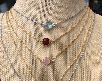 May include: Five delicate necklaces with different colored gemstones. The necklaces are layered on a mannequin's neck. The gemstones are aquamarine, red, rose quartz, amethyst, and teal.