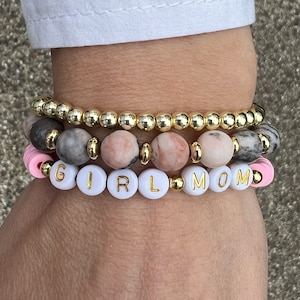 May include: A stack of three bracelets. The bottom bracelet is made of gold beads. The middle bracelet is made of pink and gray marble beads. The top bracelet is made of white beads with the word "GIRL MOM" spelled out in gold letters.