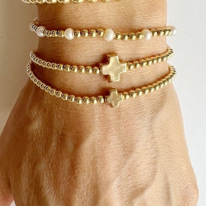 May include: Three gold beaded bracelets with cross charms. The top bracelet features white pearl beads.
